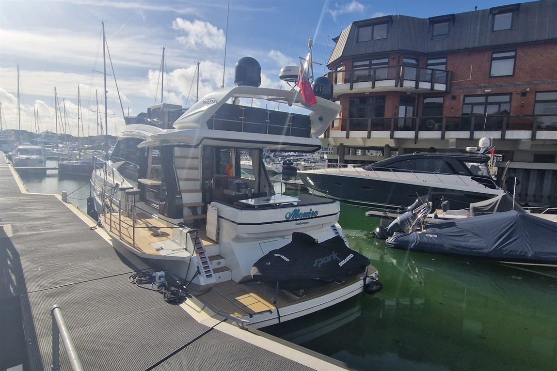 A luxury yacht named "Mannie" docked at a marina, surrounded by other boats and a waterfront building in bright sunlight.